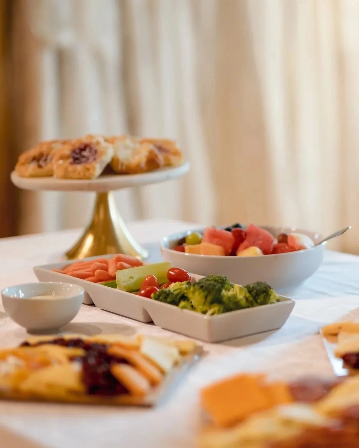 A table with a variety of colorful foods, including a bowl of fruit salad, a platter of vegetables, and a tiered stand with cookies or pastries against a blurred background.
