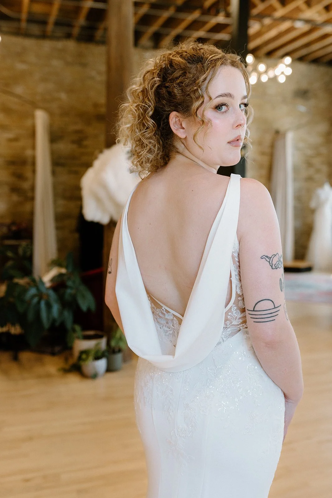 A woman with curly hair wearing a white dress with lace details, standing indoors in a rustic setting with wooden beams, plants, and decorative lighting in the background, looking over her shoulder.