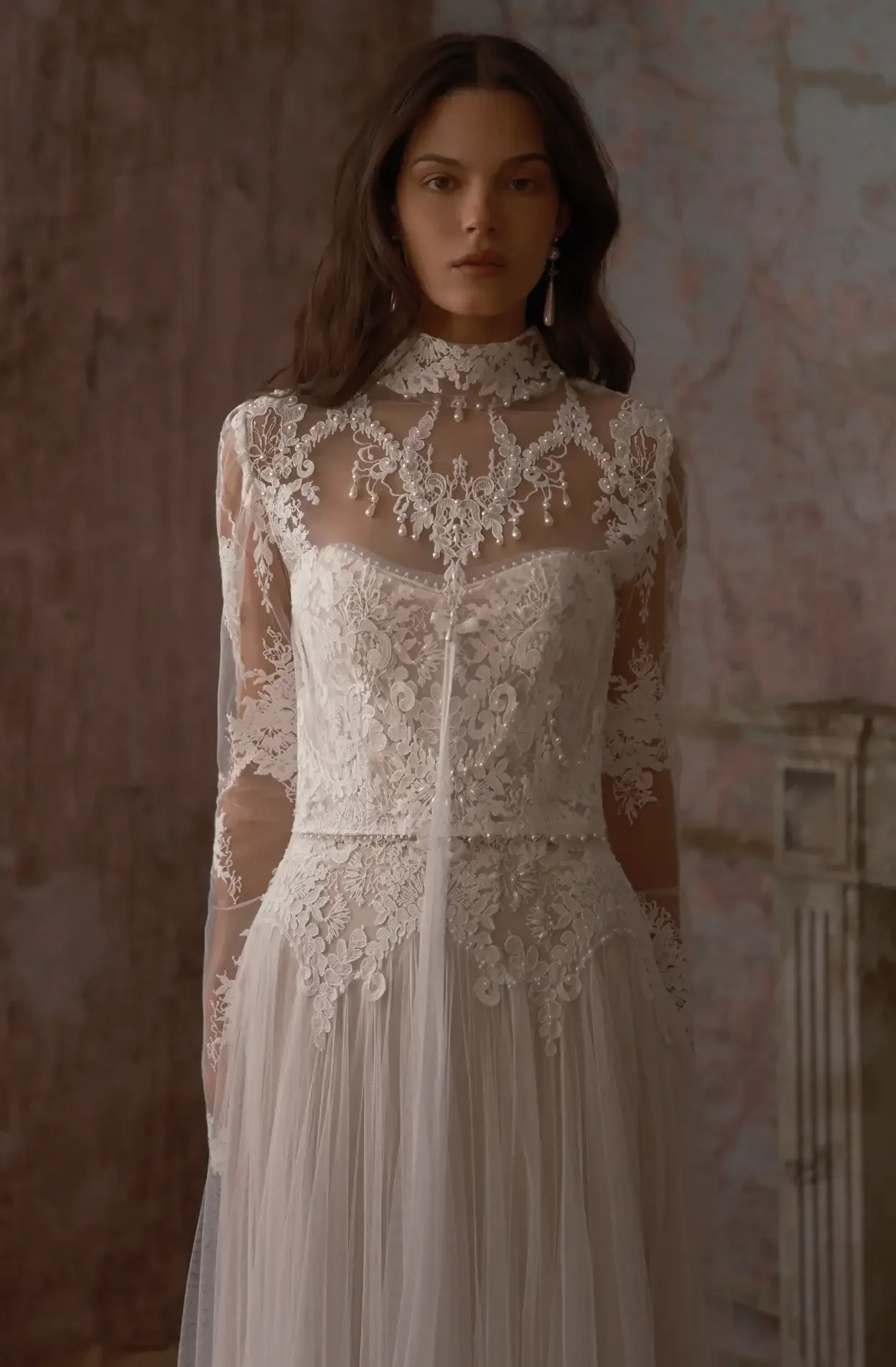 A woman in an elegant white lace wedding dress with intricate embroidery, pearl details, and a sheer overlay, standing indoors against a textured wall.
