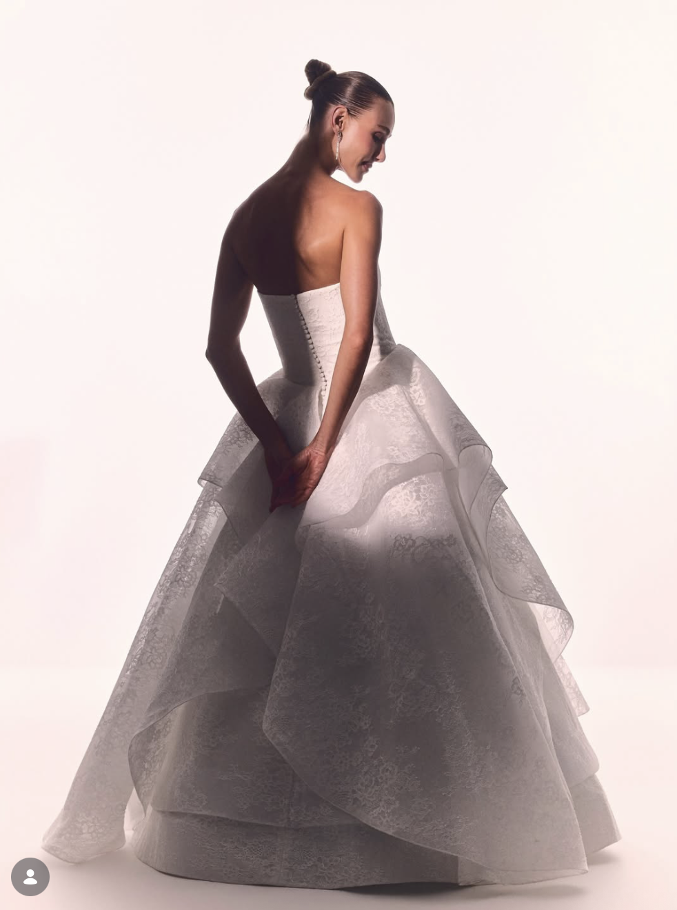 A woman in a strapless, white, lace wedding gown with layered skirt posing against a plain background.