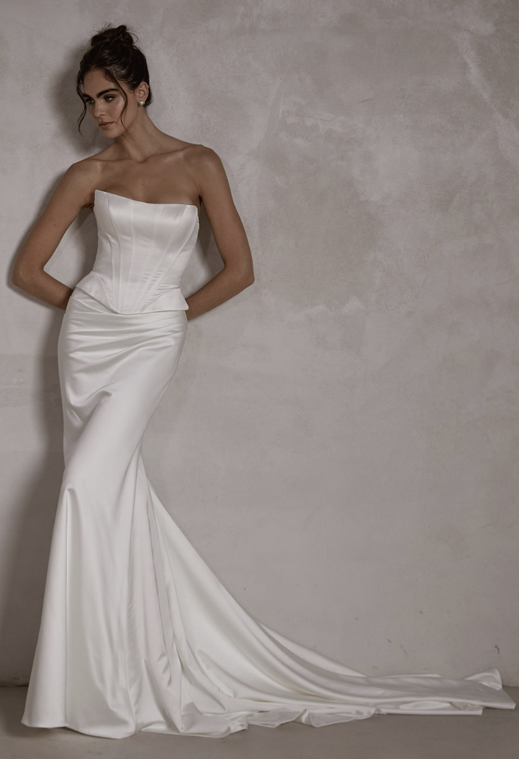 A woman in a white wedding dress leaning against a plain concrete wall.