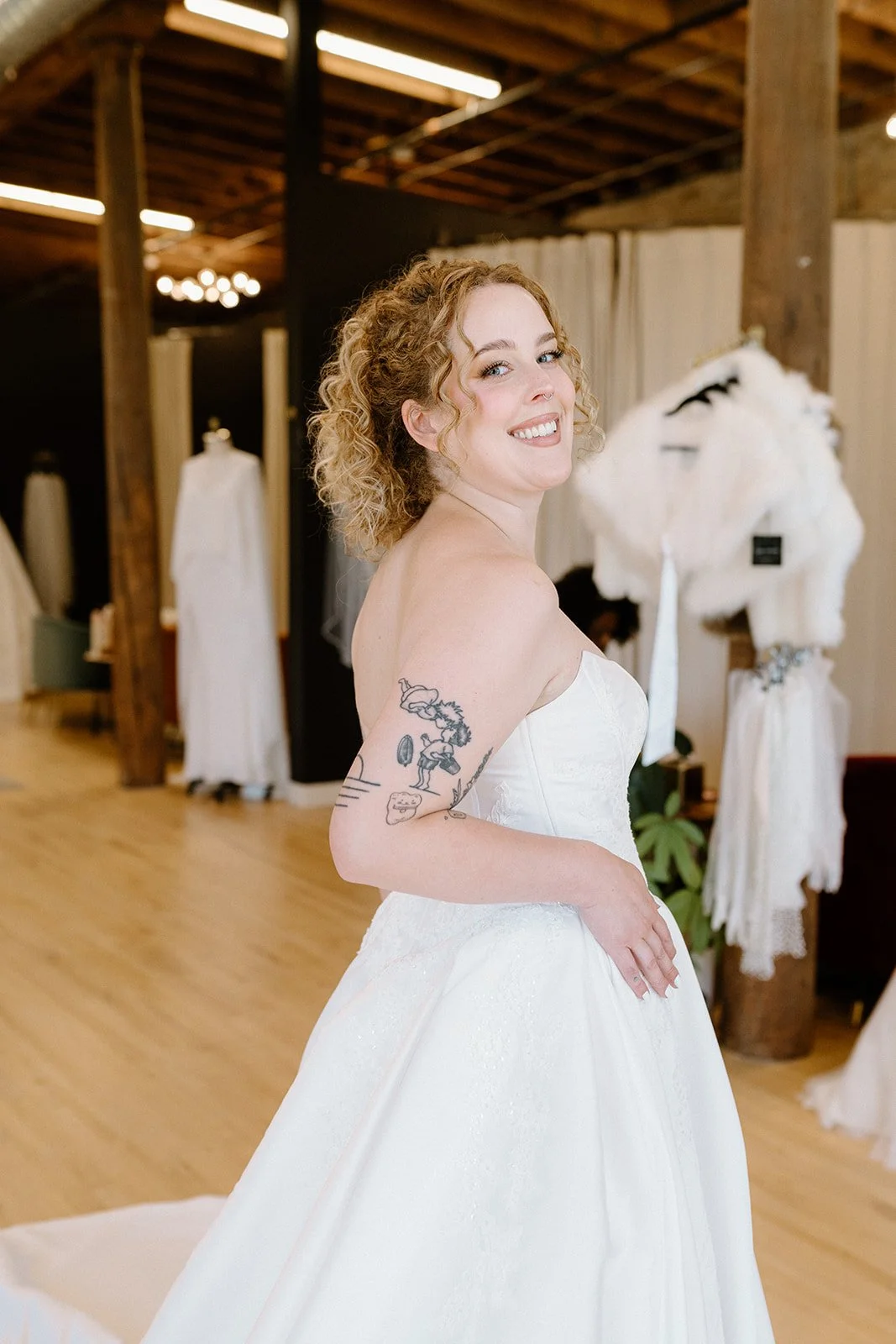 Woman in wedding dress smiling indoors with wooden beams, mannequins, and draped fabric background.