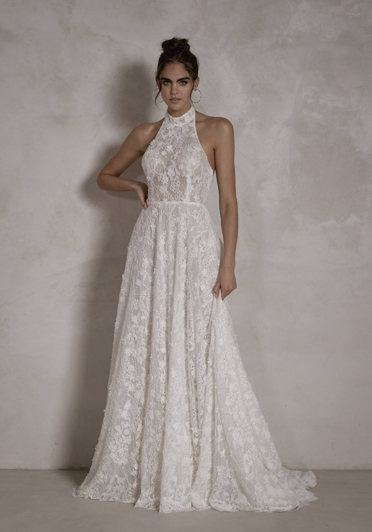 Woman modeling a sleeveless, high-neck white lace wedding dress with a fitted bodice and long flowing skirt, standing against a plain textured wall.