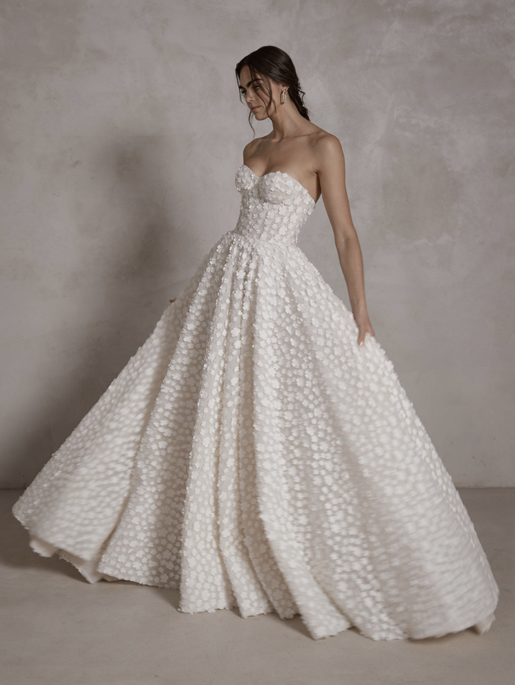 A woman in an elegant white strapless wedding gown with textured floral details, standing against a plain background.
