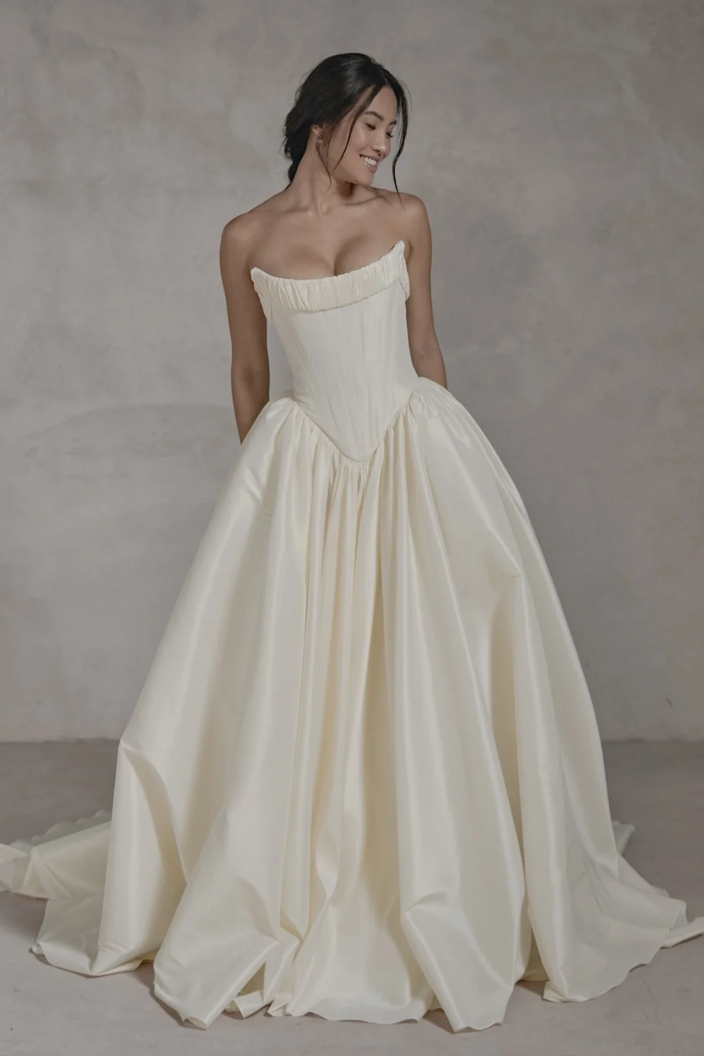 A woman in a strapless ivory satin ball gown with a fitted bodice and full skirt, standing against a plain neutral background.