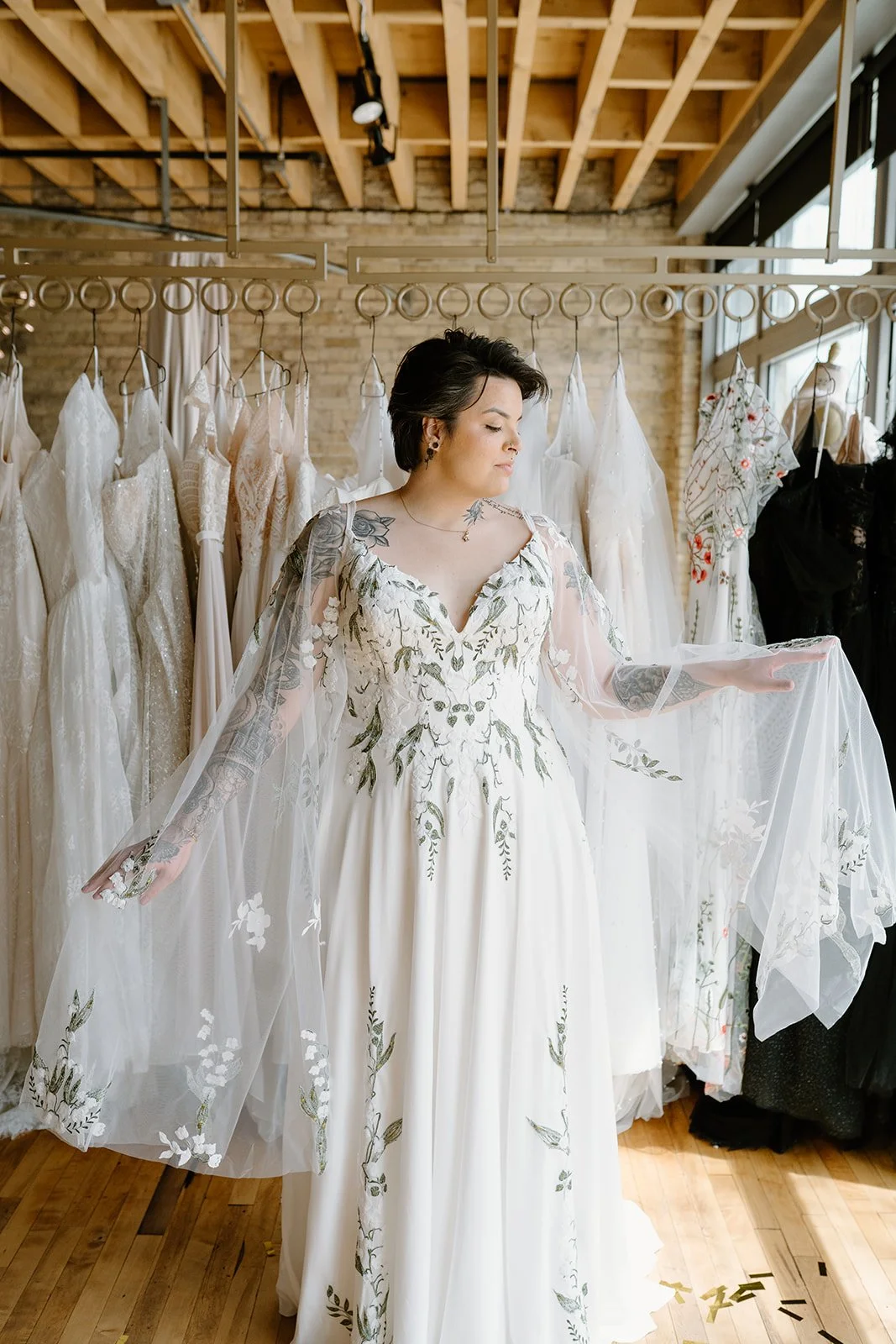 A woman in a wedding dress with floral embroidery stands in a bridal boutique, surrounded by other wedding dresses on hangers.