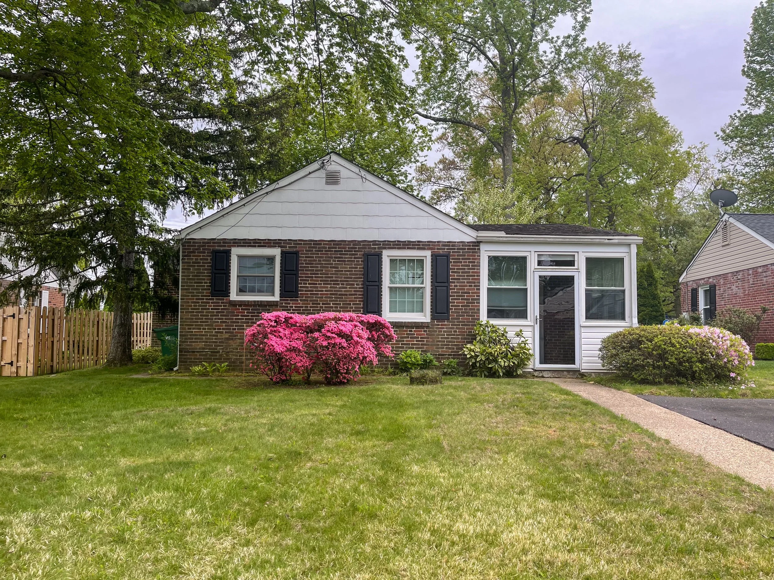 A suburban house with brick walls and white siding, surrounded by a well-kept lawn, pink flowering bushes, and tall green trees, with a concrete driveway and a satellite dish on the roof.