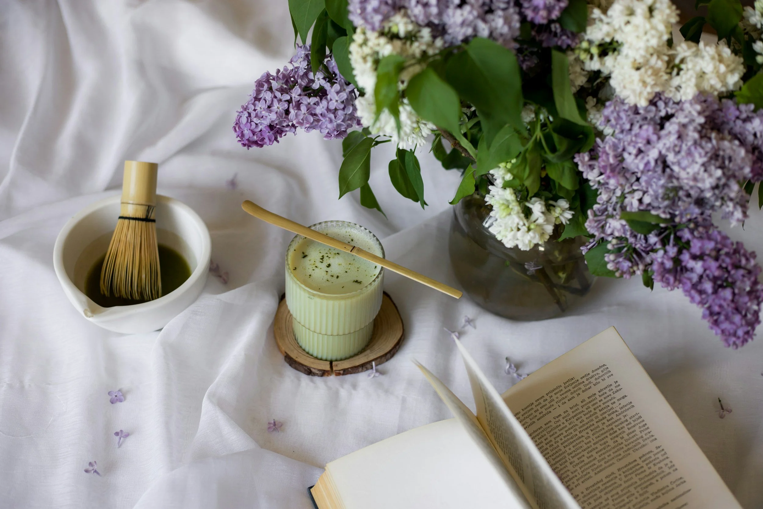 Soft image of flowers, a book, and tea on white sheets evokes mindfulness, rest, and self-compassion as antidotes to perfectionism and stress.