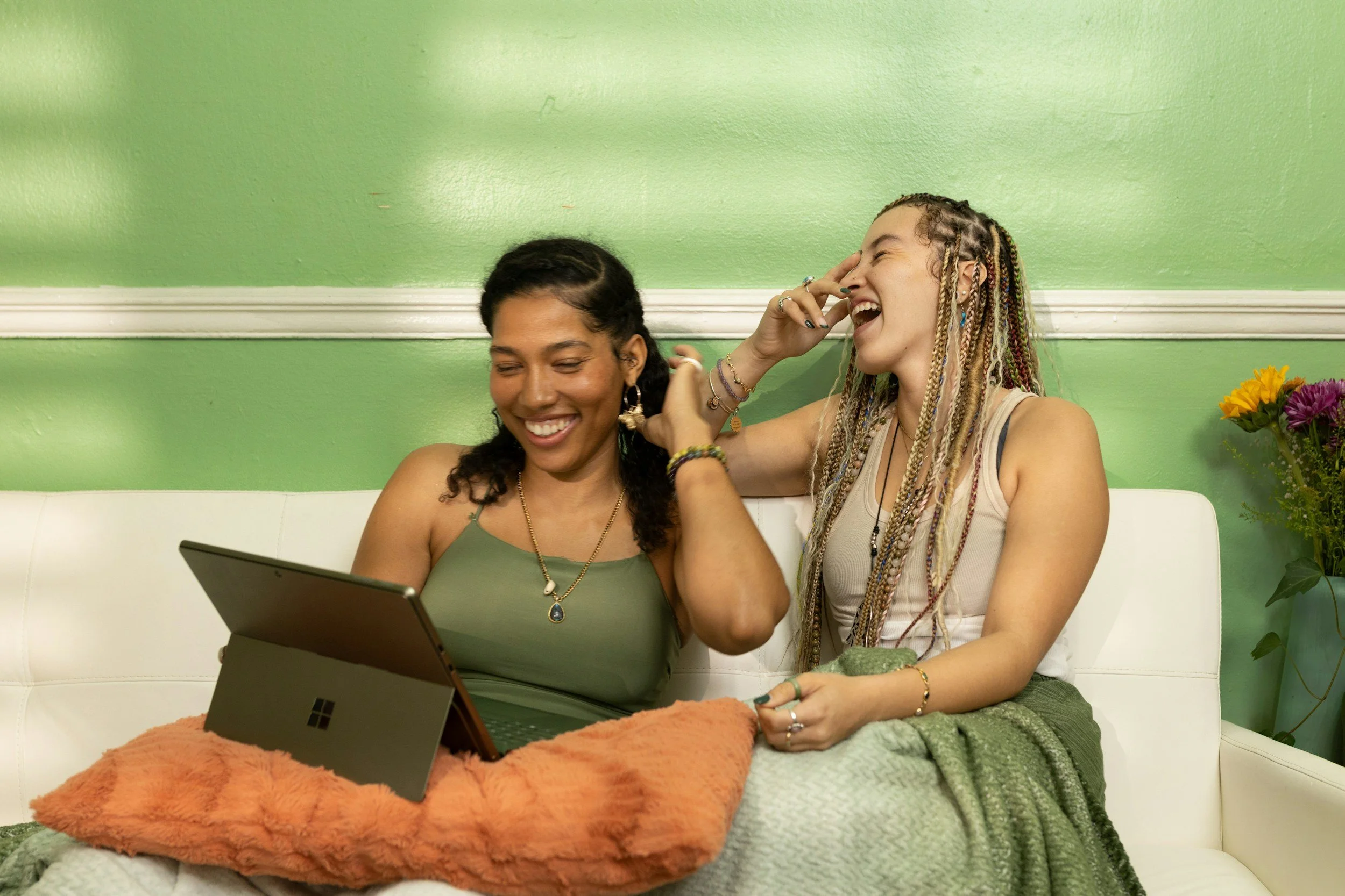 Two women sitting together on a couch, smiling and laughing while using a laptop, representing supportive collaboration and how shared systems and technology reduce mental load and perfectionism in OCD recovery.