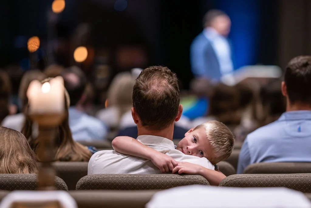 A father and young boy are seated in the worship center, with the boy resting his head on the father's shoulder.