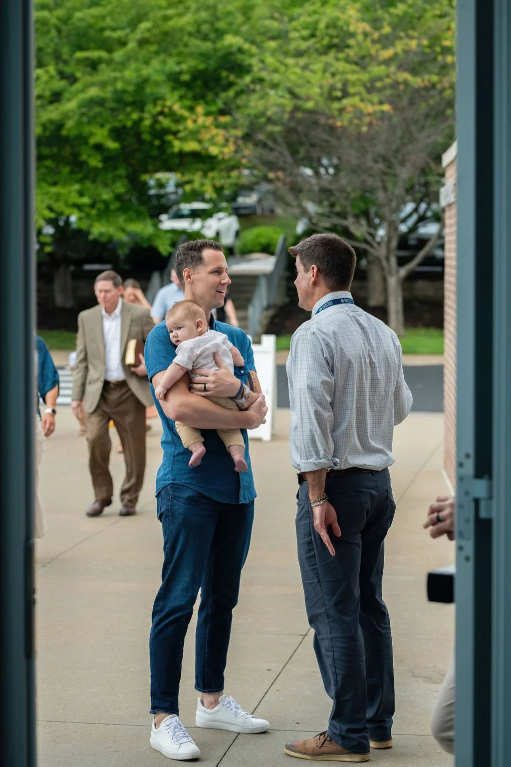 Two men engaged in conversation outside the worship center, one is holding a baby, and a few people are walking in the background, with trees and cars visible.