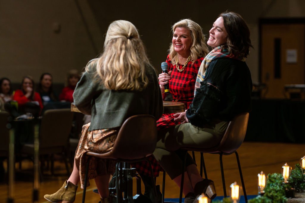 Three women sitting on chairs having a conversation at a Women's ministry event with audience in the background. One woman, holding a microphone, smiles as she talks to the others.
