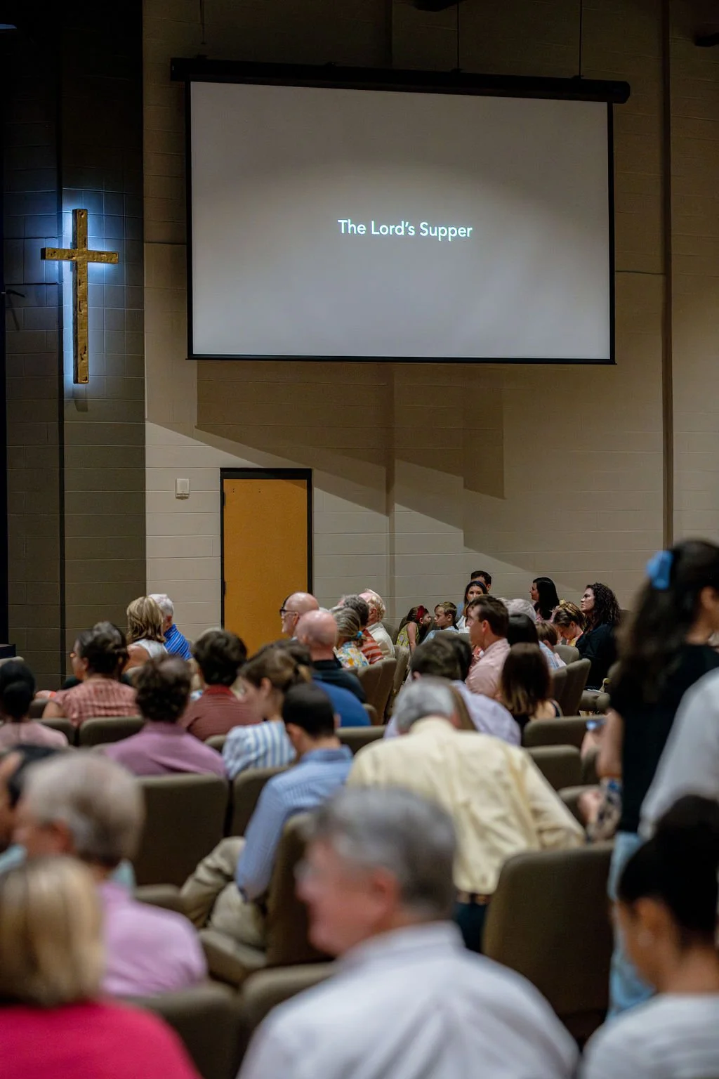 A church congregation seated in the worship center, watching a presentation on a large screen that reads 'The Lord's Supper.' A wooden cross is mounted on the wall to the left of the screen.