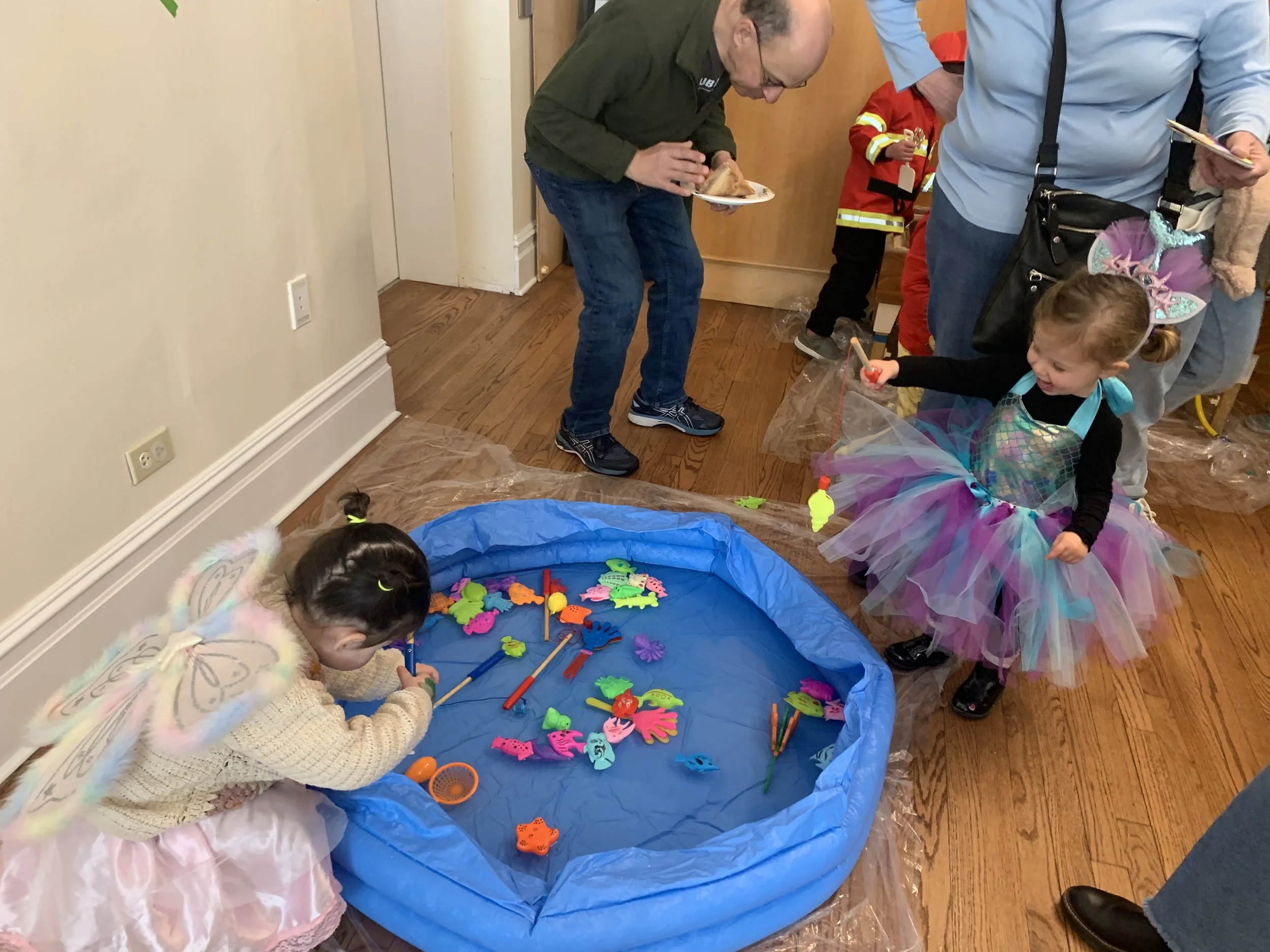 two preschool-aged girls dressed in purim fairy costumes play with toys in a kiddie pool at the synagogue