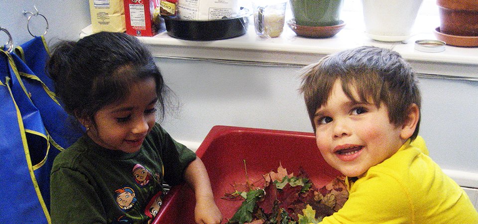 two smiling preschool children play with leaves in class