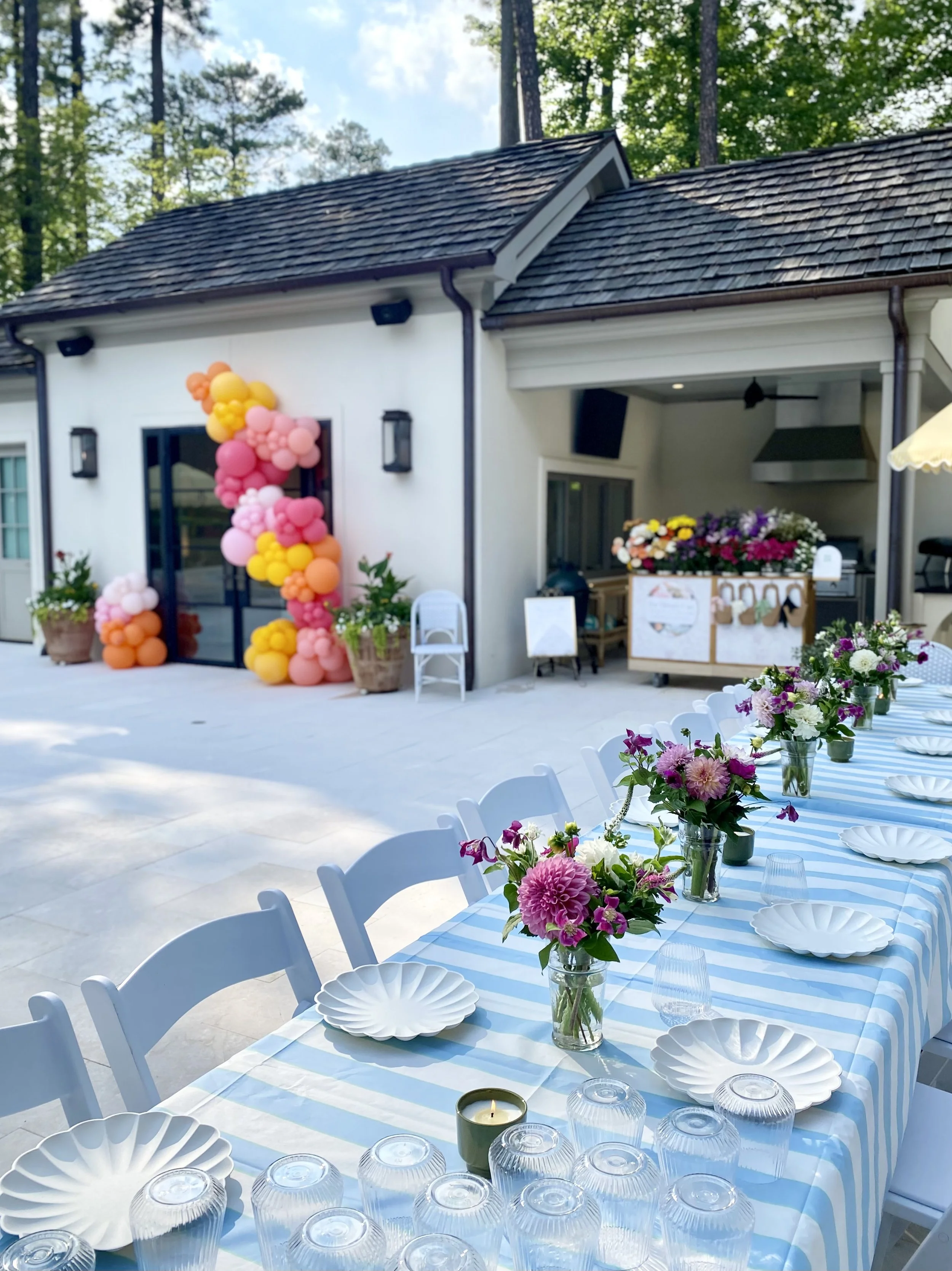 a tablescape on an outdoor patio with flowers and a flower bar in the background and a colorful balloon arch