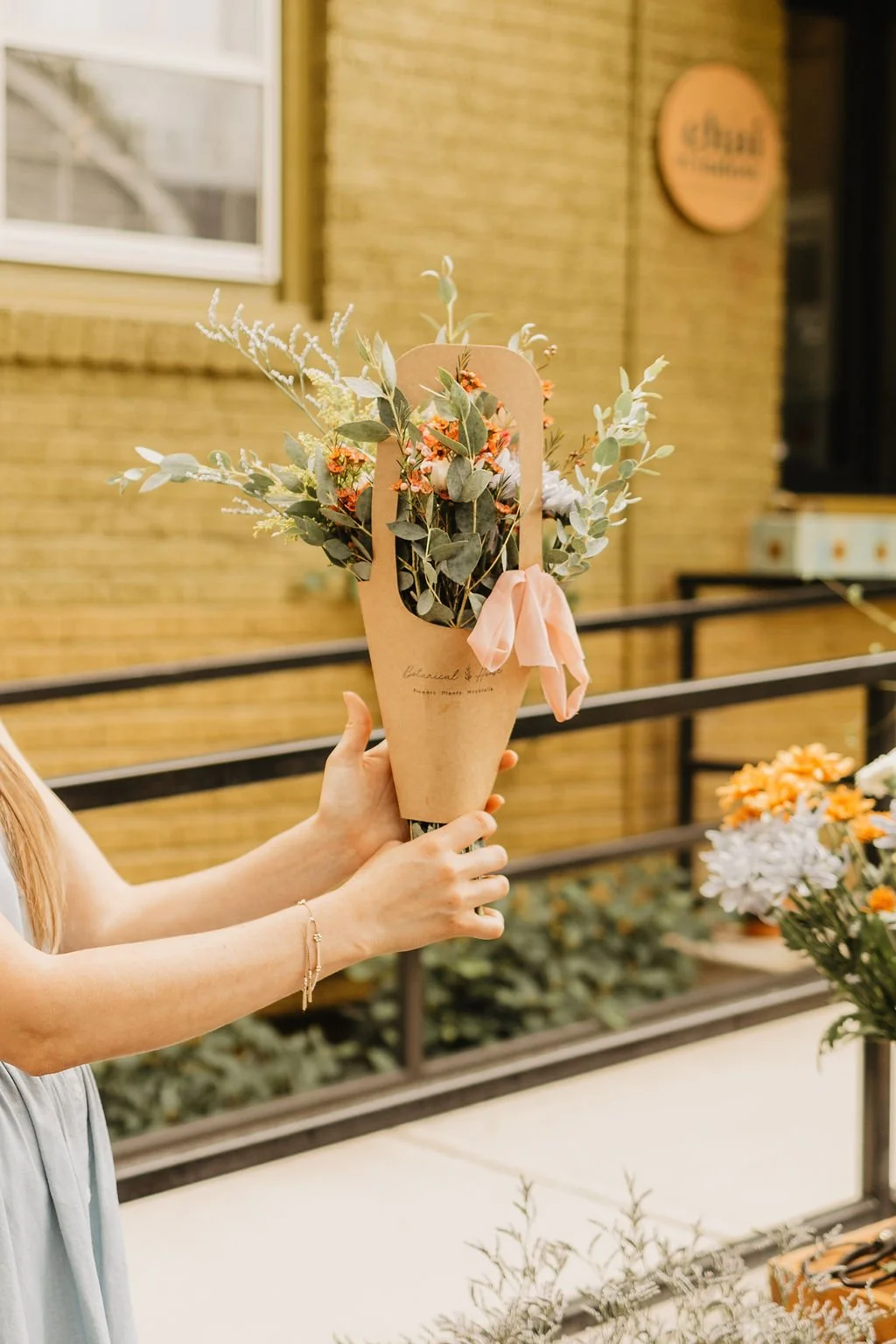 a bouquet of flowers in a brown paper carrier with a pink bow being held by two hands outside in front of a green brick building