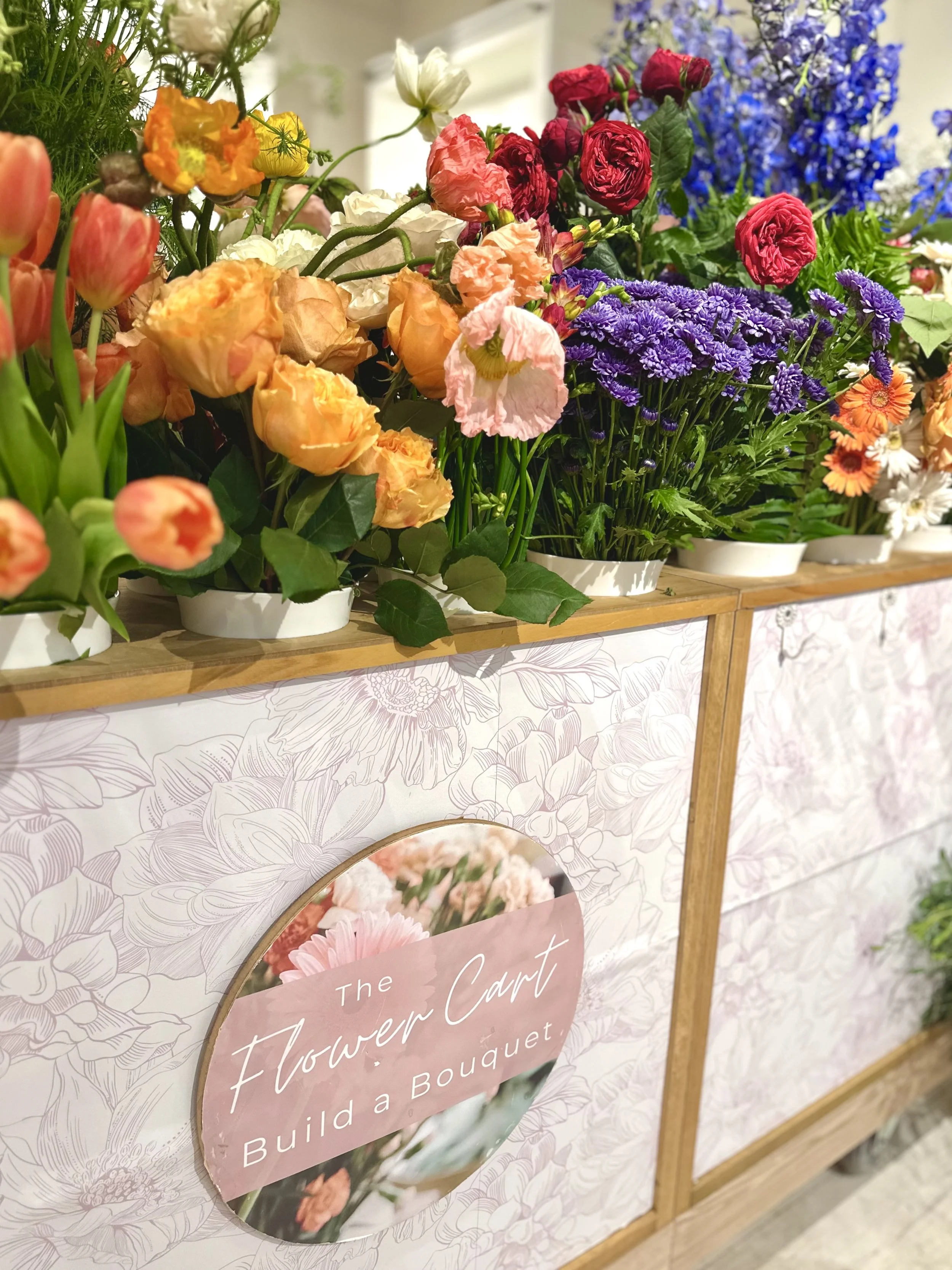 A flower cart that is stocked with bright fresh flowers and has a sign that says “the flower cart, build a bouquet”