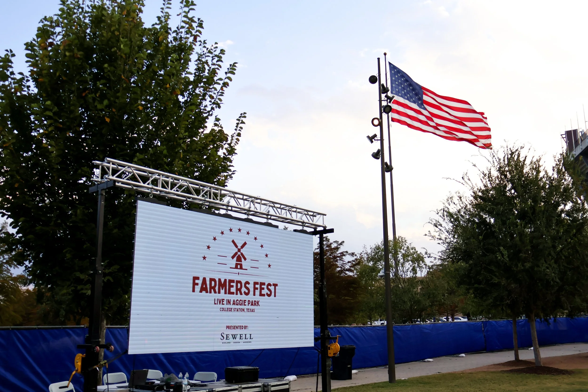 An outdoor scene at a Farmers Fest event at Aggie Park, College Station, Texas. There is a large white event banner with the text "FARMERS FEST LIVE IN AGGIE PARK COLLEGE STATION, TEXAS PRESENTED BY: SEWELL" hanging on a frame. A flag of the United States is flying on a pole nearby. Trees and a partly cloudy sky are in the background.