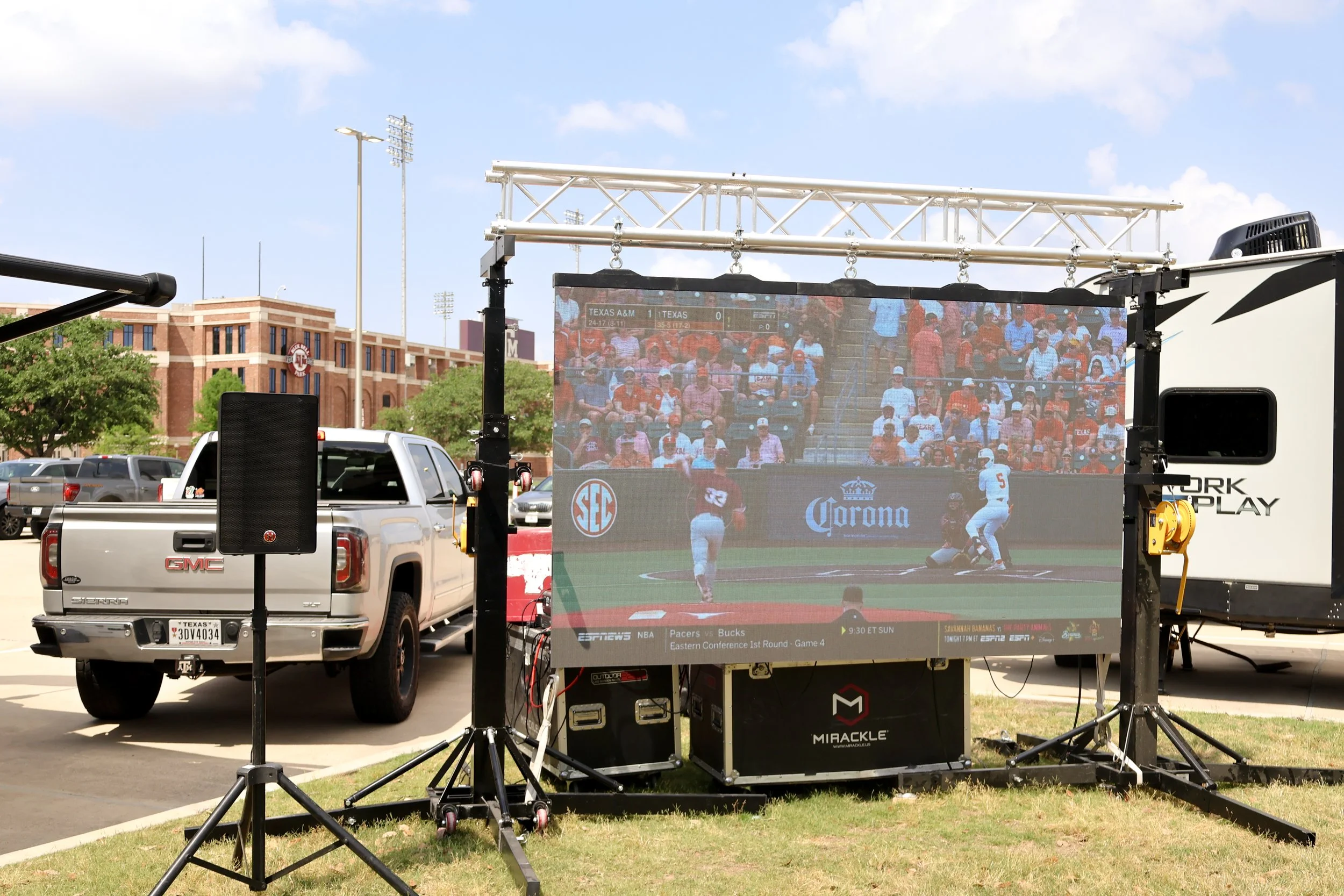 Outdoor setup with large screen showing a baseball game, surrounded by trucks and equipment, in a parking lot near a stadium.