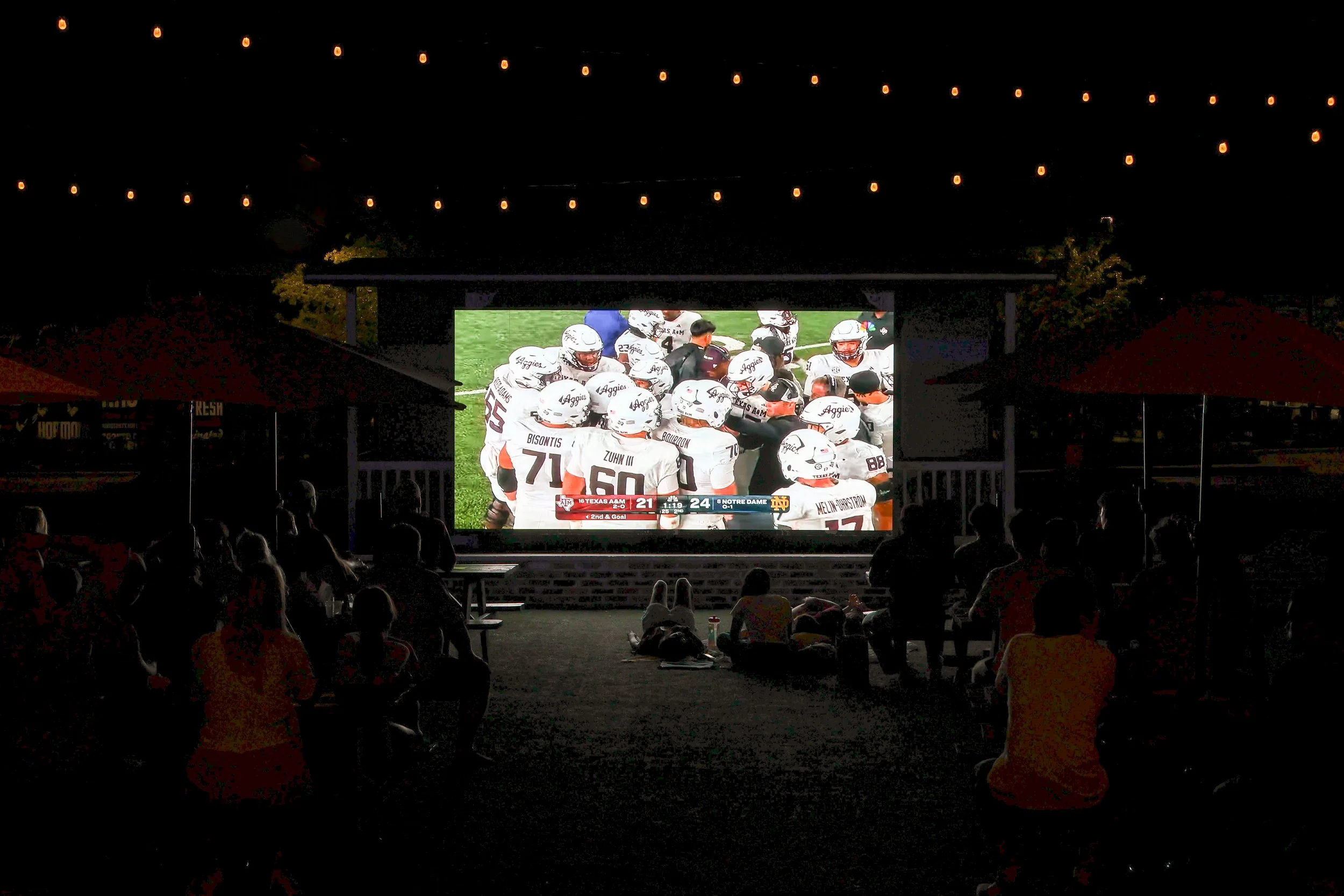 People sitting outdoors at night, watching a football game on a large outdoor screen, with string lights overhead.
