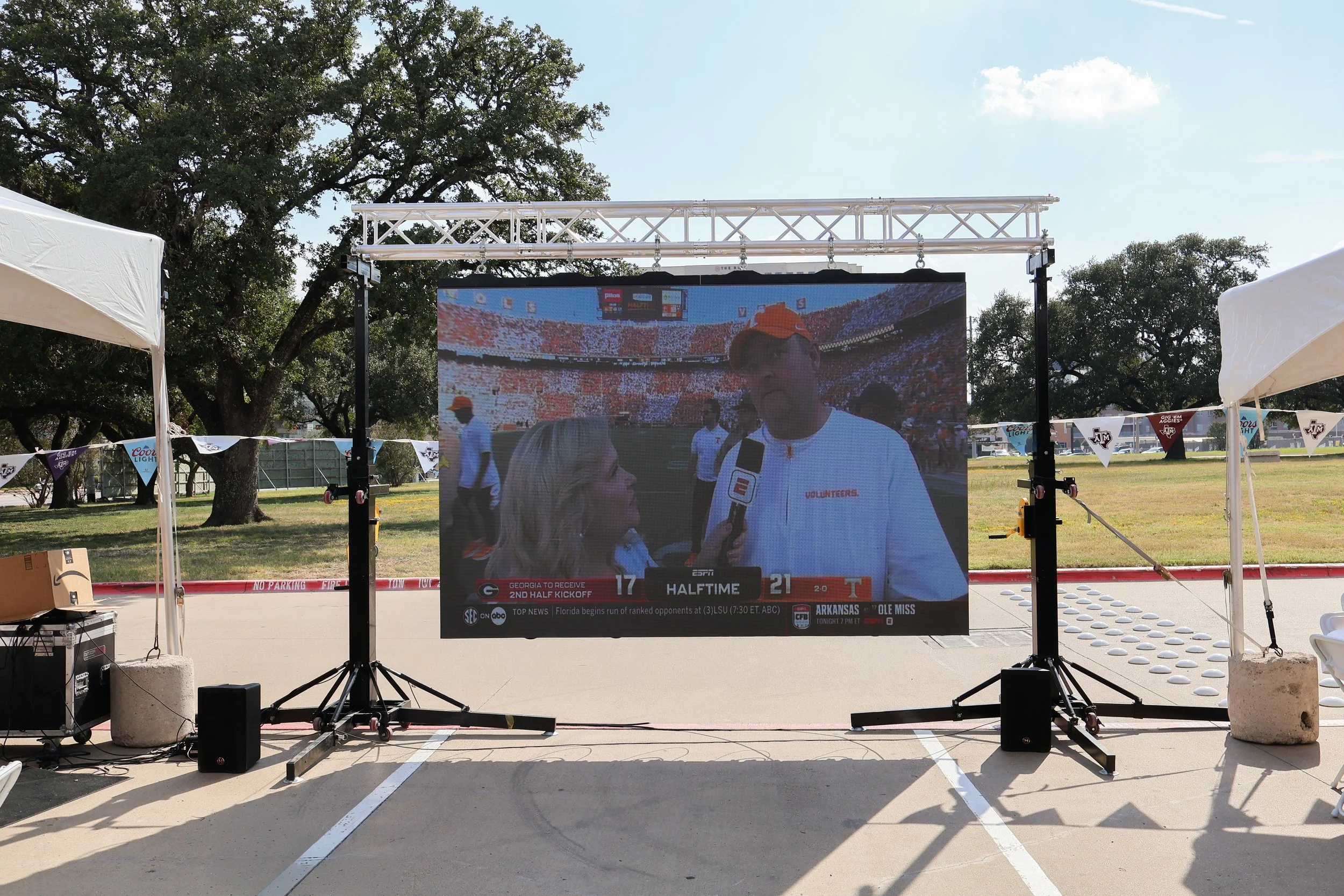 An outdoor large digital screen displaying a football interview with a man in a white shirt and orange cap, holding a microphone, in a stadium filled with spectators, with some sponsorship logos and a scoreboard visible.