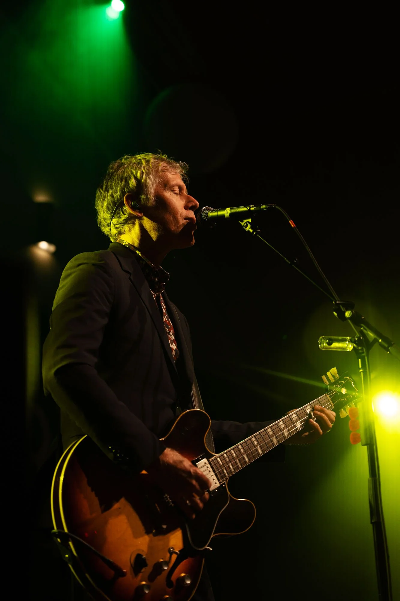 A man performing on stage with a guitar, singing into a microphone, illuminated by green and yellow stage lights.