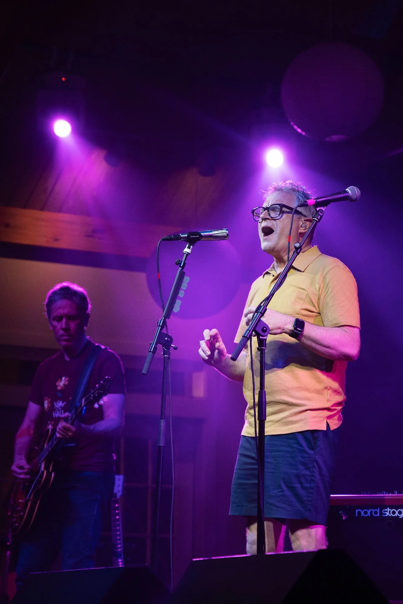 A man in yellow shirt and glasses singing into a microphone on stage with purple lighting, accompanied by a guitarist.