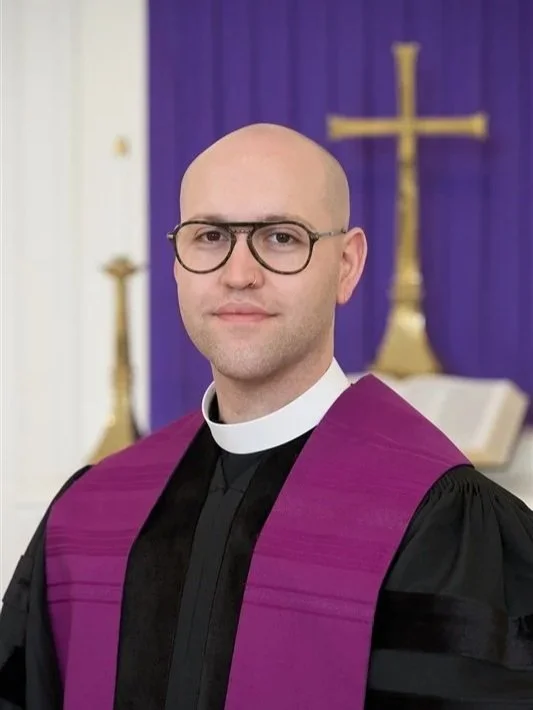 A smiling man with glasses and a closely shaved head, wearing a clerical collar and black shirt, standing against a light-colored wall.