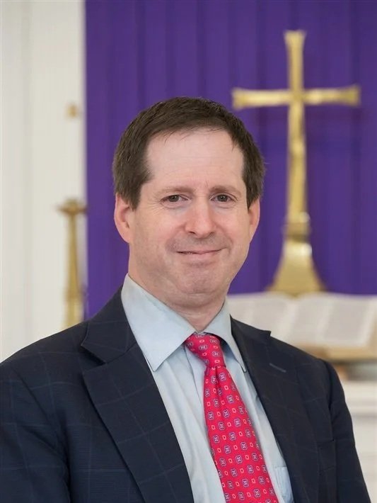 A man in a suit and red tie smiling in front of a chancel with a gold cross and purple fabric.