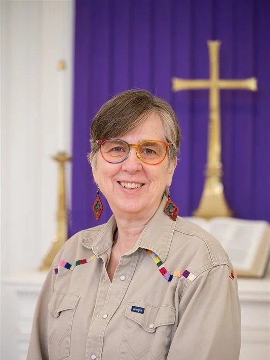 Portrait of a smiling woman wearing glasses, earrings, and a tan collared shirt in front of a chancel with a gold cross and purple fabric.