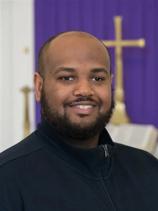 A man with a black beard and a smiling expression in a black sweater in front of a chancel with a gold cross and purple fabric.