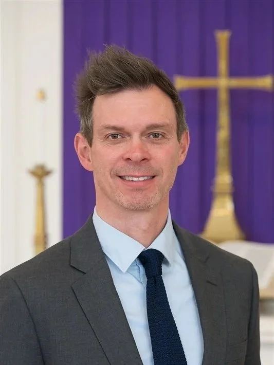 A smiling man in a suit and collared shirt standing indoors in front of a chancel with a gold cross and purple fabric 