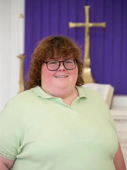 Portrait of a smiling woman wearing glasses and a light green collared shirt in front of a chancel with a gold cross and purple fabric.