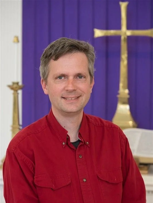 Portrait of a smiling man in a red collared shirt in front of a chancel with a gold cross and purple fabric.