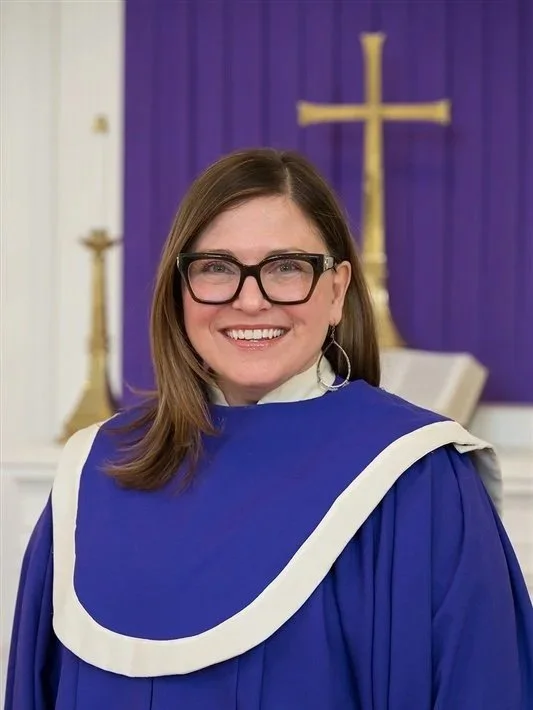 A woman smiling, wearing large tortoiseshell glasses and a purple choir robe, standing indoors in front of a chancel with a gold cross and purple fabric.