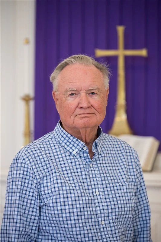 Portrait of an older man with grey hair and a serious expression in a blue collared shirt standing indoors in front of a chancel with a cross and purple fabric.