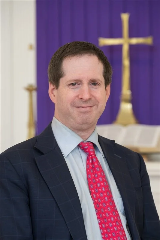 A man in a suit with a red tie, smiling, standing in front of a chancel cross.