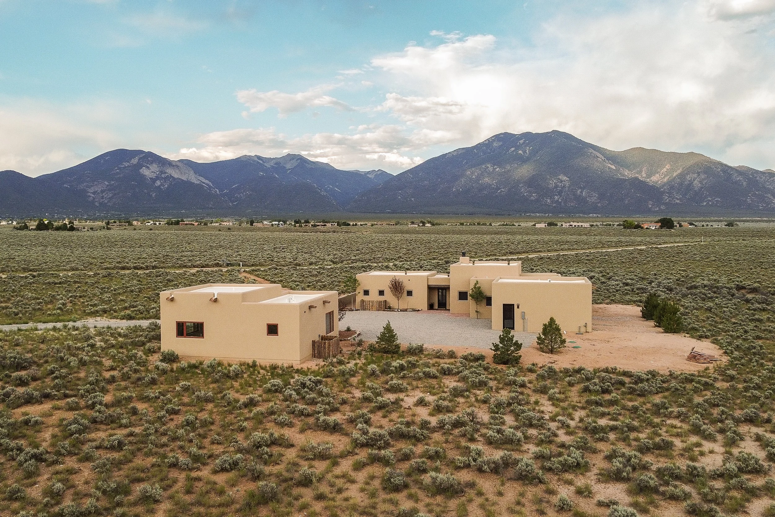 A modern house with beige walls and flat roof in a desert landscape with scattered shrubs and small trees, mountains in the background under a partly cloudy sky.