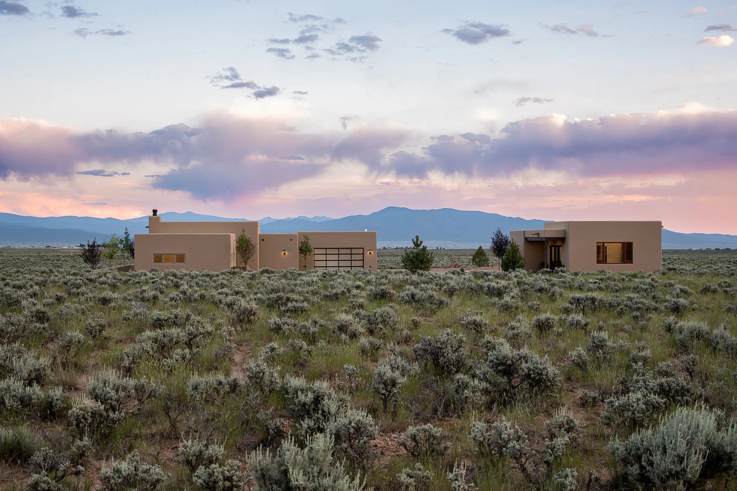 Modern beige houses in a desert landscape with low shrubs, mountains in the background, and a colorful sky with clouds at sunset.