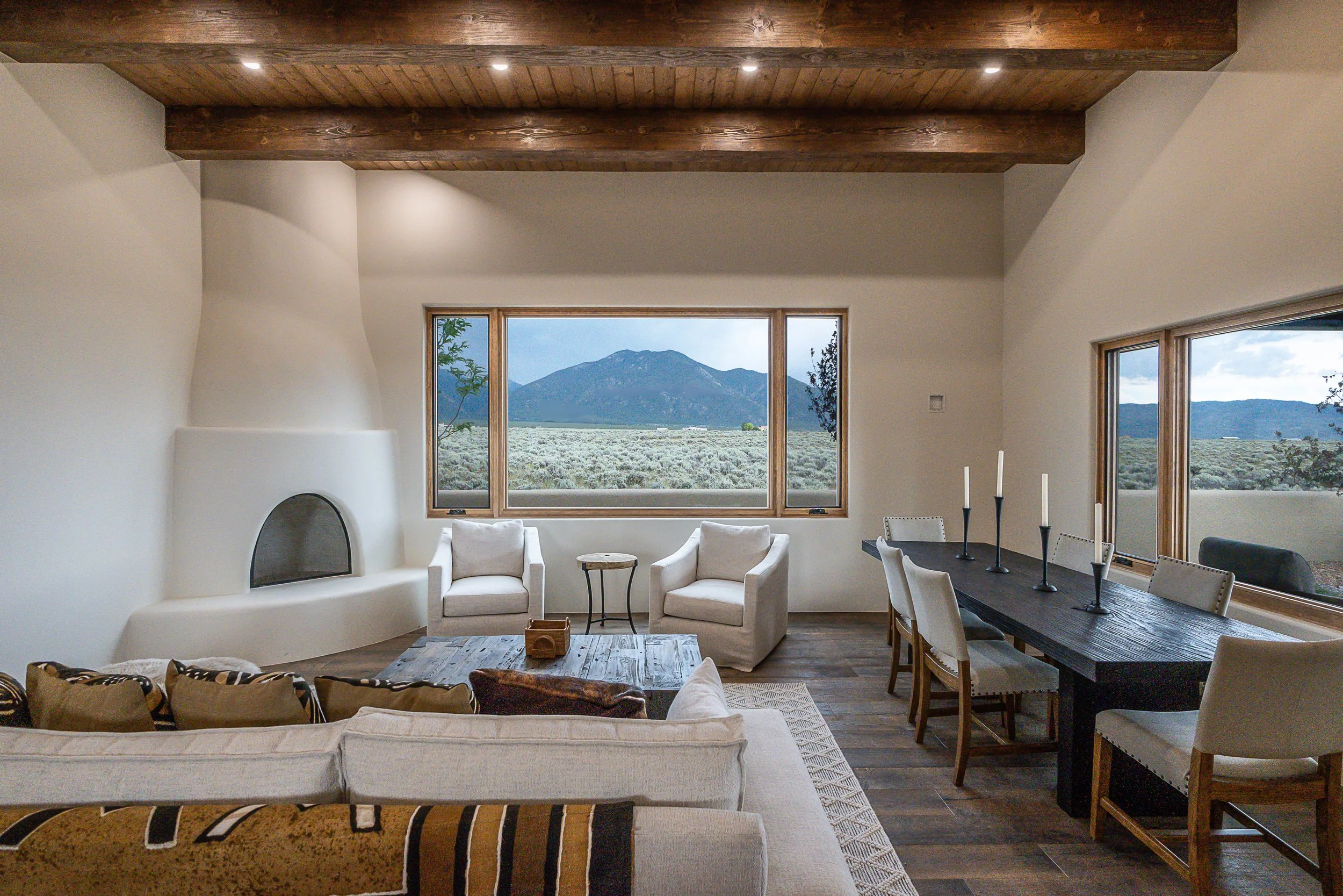 Living room with wooden beams, white walls, large window overlooking mountains, and a mix of modern and rustic furniture including a beige sofa, white armchairs, a black dining table with white chairs, and decorative candles.