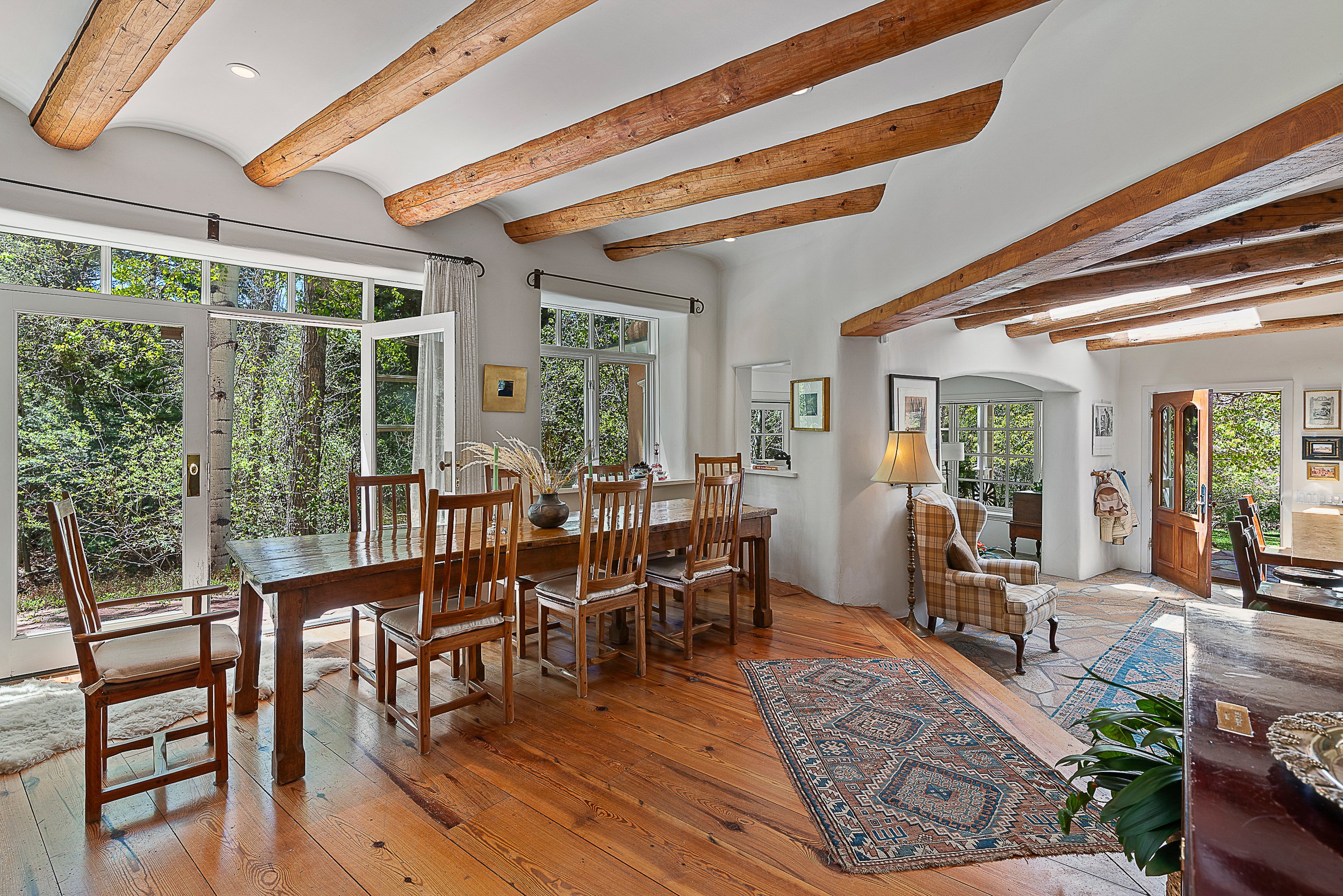 Cozy dining and living room with wooden beams, hardwood floors, and large windows overlooking trees.