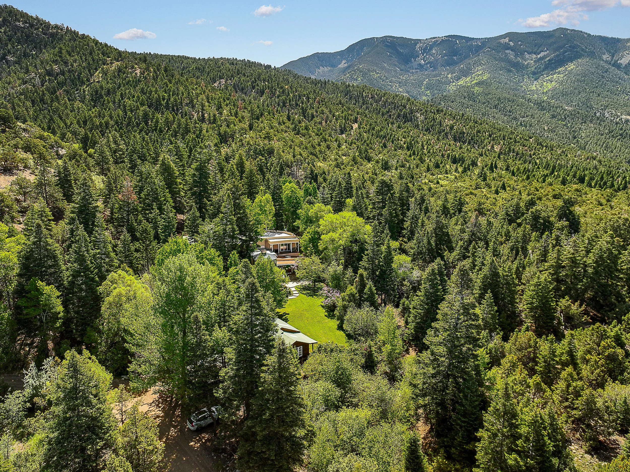 Aerial view of a house surrounded by dense green forest on a hillside, with mountains and a blue sky in the background.