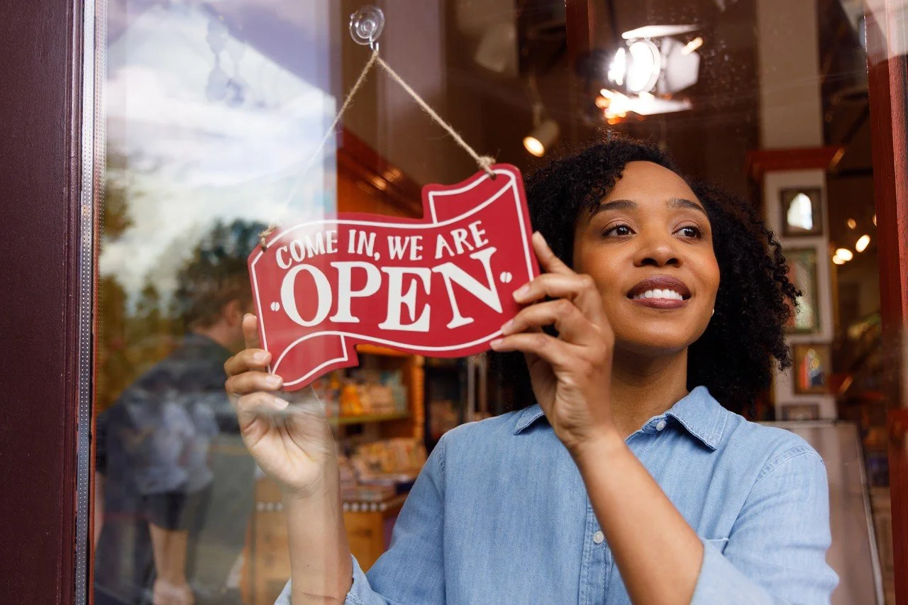 A woman with curly hair hanging an 'OPEN' sign on a shop window. She is smiling and wearing a light blue shirt. The shop interior is visible behind her.