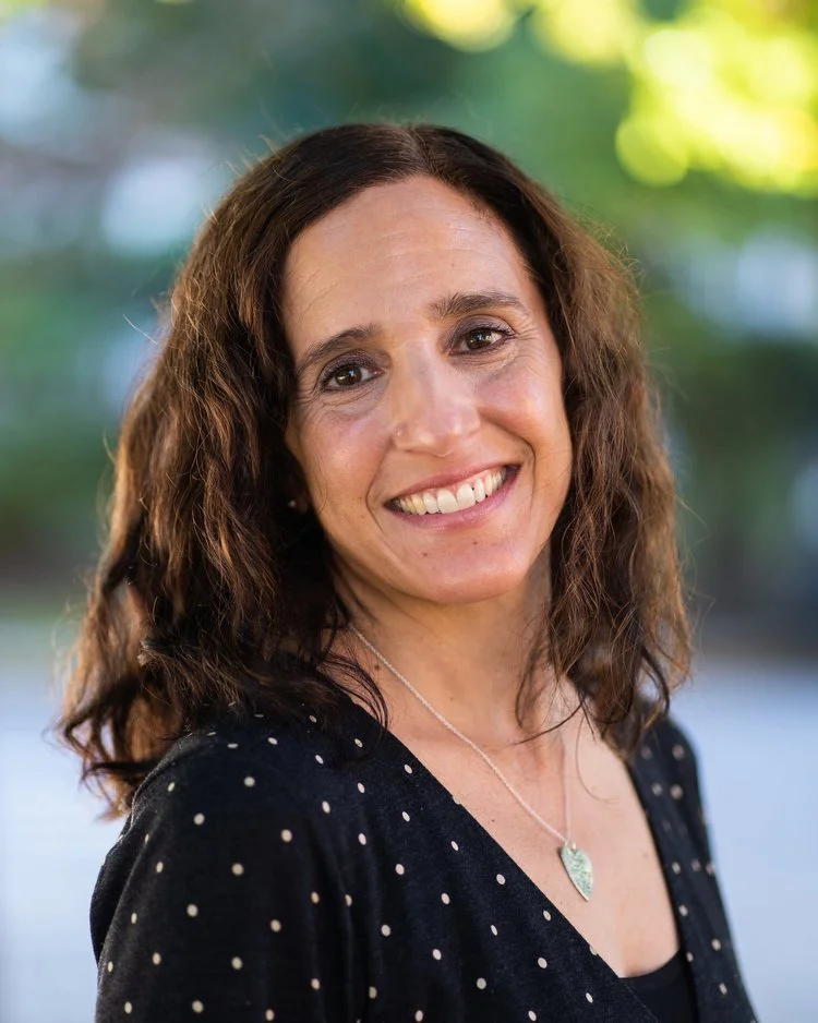 A woman with shoulder-length curly brown hair, smiling outdoors with a blurred green background, wearing a black top with white polka dots and a silver necklace.