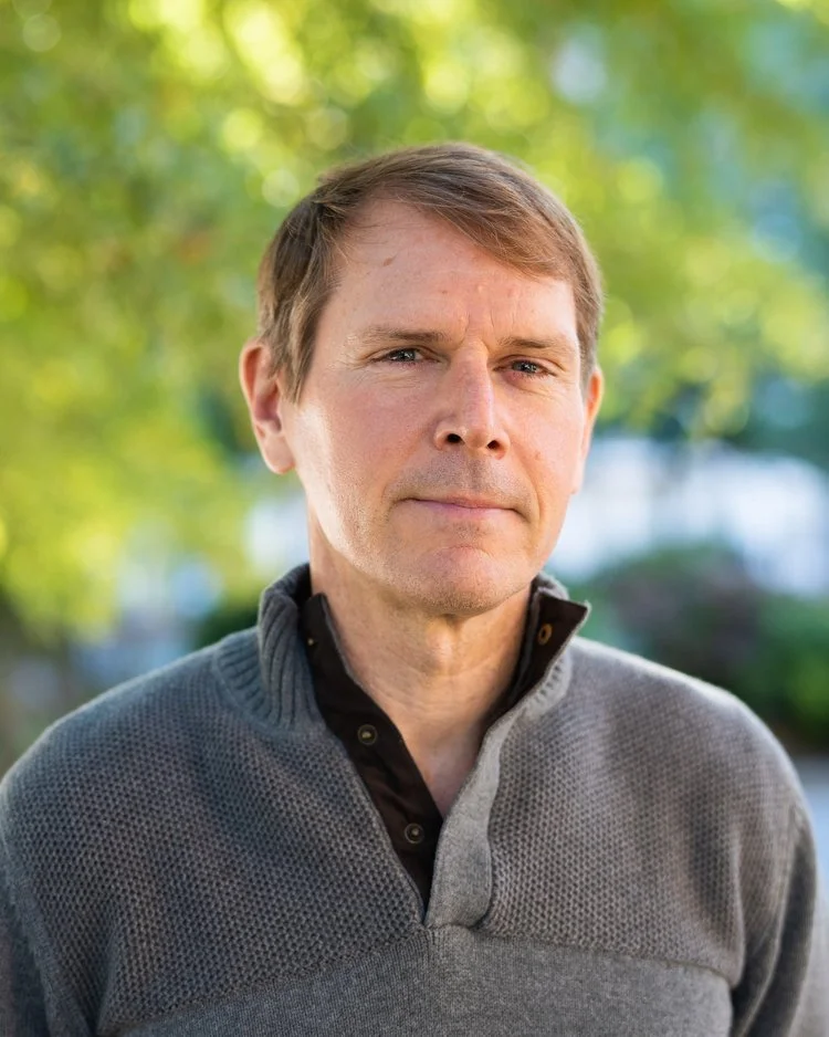 A middle-aged man with light brown hair, wearing a gray sweater over a black collared shirt, standing outdoors with blurred green trees in the background.
