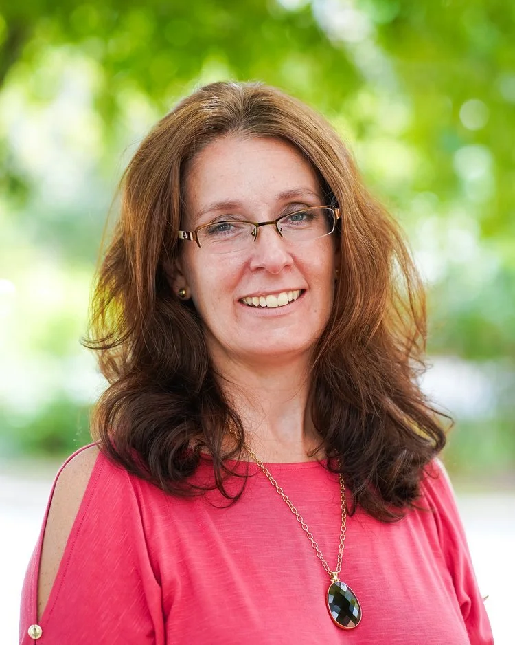 A woman with brown hair, glasses, and a pink top, smiling outdoors with green foliage in the background.