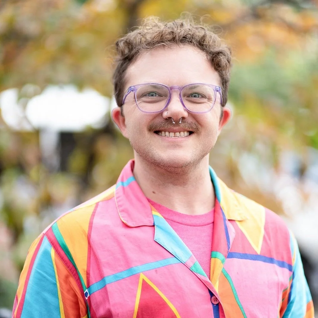 A young man smiling outdoors with blurred colorful autumn trees in the background. He is wearing clear purple glasses, a colorful striped shirt, and has a nose piercing and facial hair.