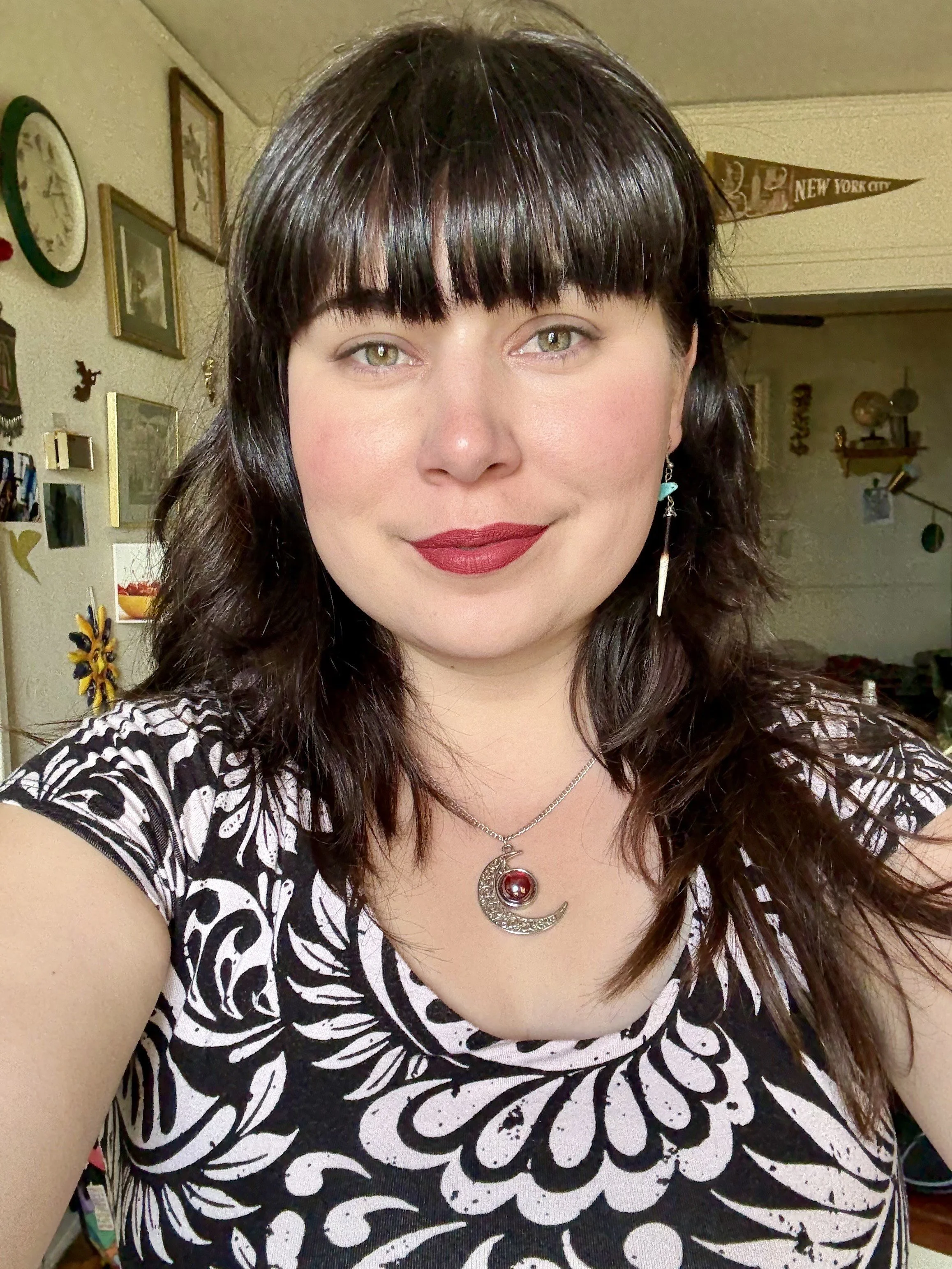 A woman with dark brown hair, bangs, and light green eyes, wearing a black and white patterned top and a moon and star necklace with a red gemstone, smiling in a room decorated with clocks, picture frames, and various wall hangings.