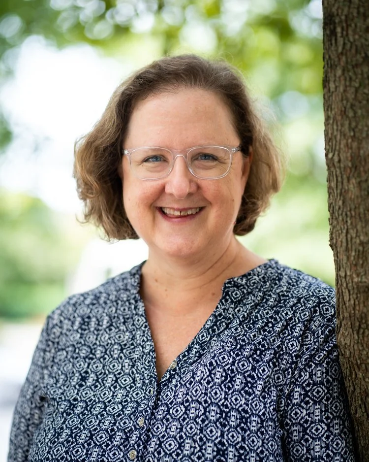 A smiling woman with glasses and short, wavy hair, standing outdoors next to a tree with a green, leafy background.